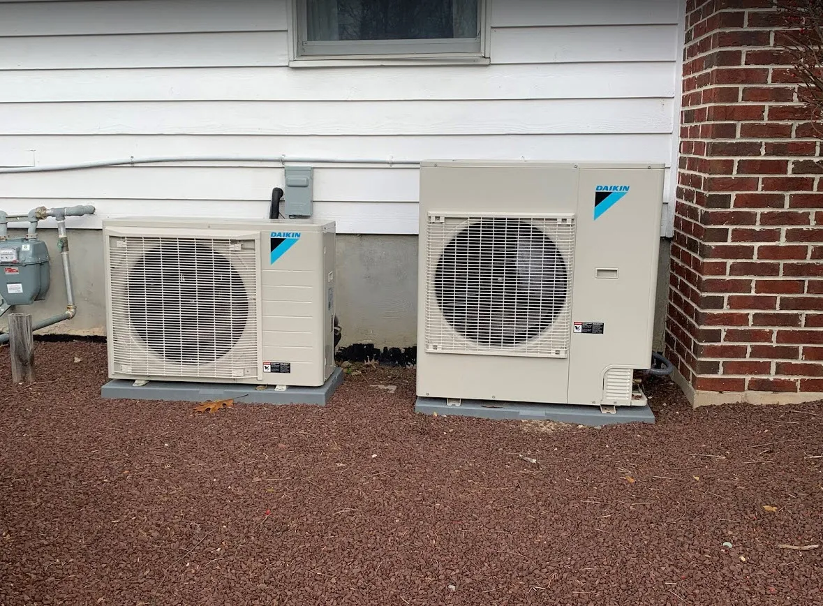 Ducltess units beside a house with white siding and a brick wall