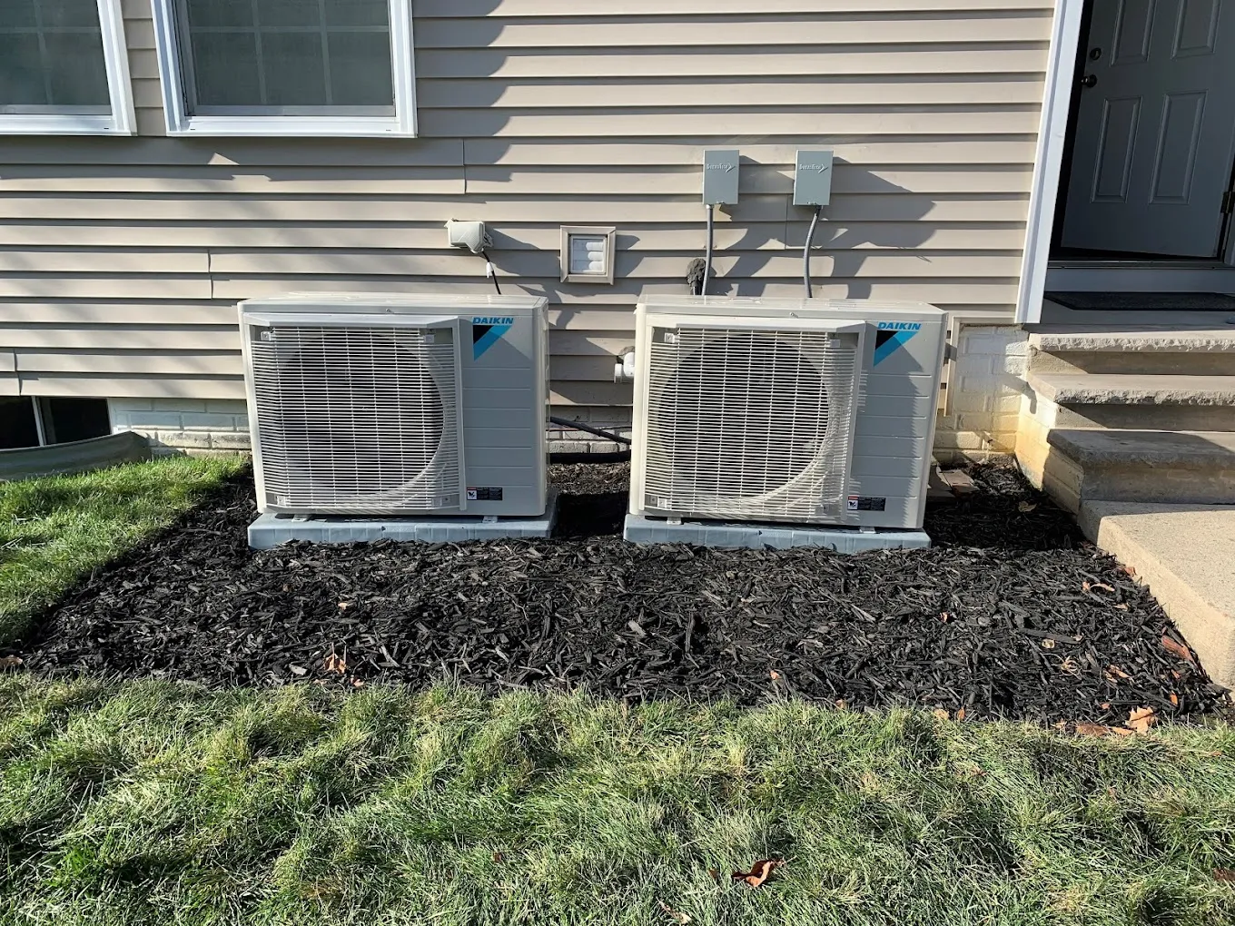 Two outdoor AC units on mulch beside a house with steps and a door